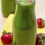 Healthy green juice smoothie in glass bottle sitting on yellow napkin with fresh strawberries.