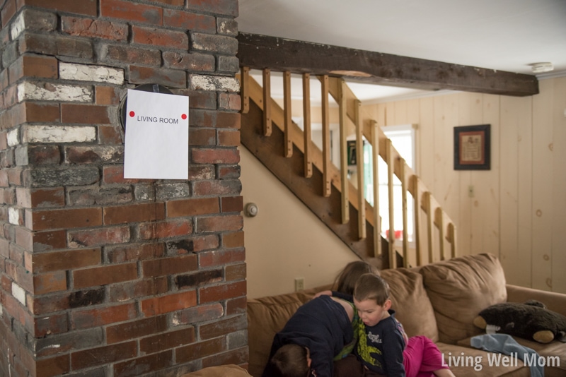 A brick wall in a living area of a house, with a paper reading "Living Room" hung on the wall, with red stickers on it. 