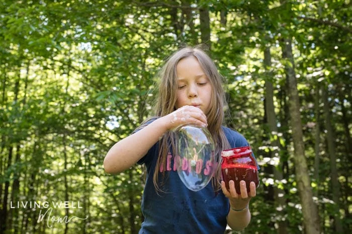 A child blowing homemade bubble solution with one hand, and a red jar of the bubbles in another. 