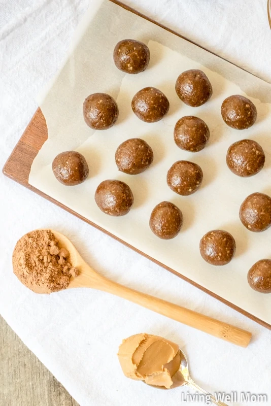 Gluten-free protein ball mixture rolled up into uniform balls on a cutting board, with spoons of almond butter and cacao powder nearby.