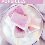 Four homemade berries and cream popsicles piled on a white plate with ice cubes, showing a soft pink and lavender swirled color from mixed berries and milk, sweetened with maple syrup and dairy-free.