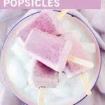 Four homemade berries and cream popsicles piled on a white plate with ice cubes, showing a soft pink and lavender swirled color from mixed berries and milk, sweetened with maple syrup and dairy-free.