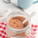 homemade cocoa mix in glass container with red checkered napkin and blue mug