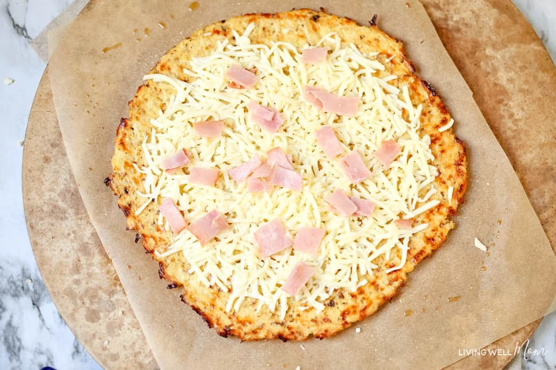Cauliflower pizza crust topped with shredded cheese, and pieces of deli ham. 