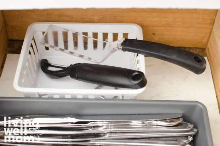 small white basket at the back of a drawer to keep utensil tray from sliding around holding a serving tool and a vegetable peeler. 