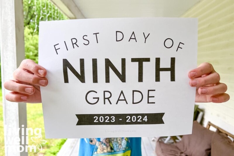 teen holding piece of paper that says first day of ninth grade 2023-2024 on porch with greenery