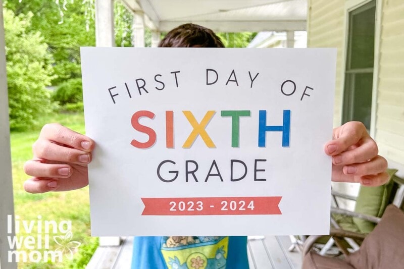 boy with blue shirt holding rainbow colored paper that says first day of sixth grade 2023-2024 