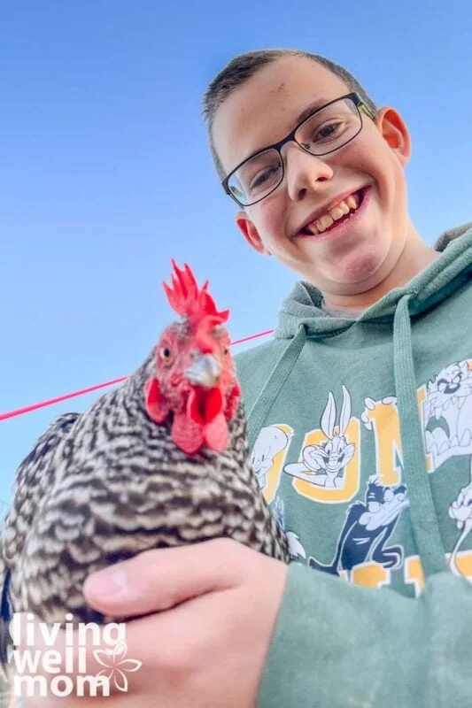 smiling boy wearing a green hoodie holding a barred rock hen chicken with blue sky