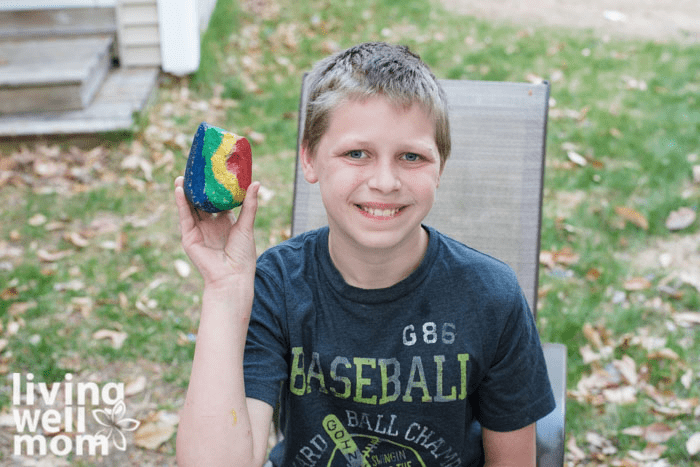 A boy holding up a rainbow designed pet rock he painted with a smile on his face. 