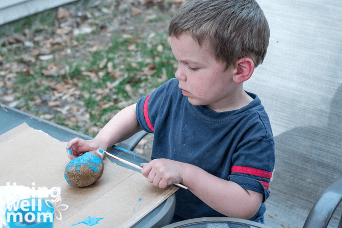 A young boy covering a round rock with bright blue paint. 