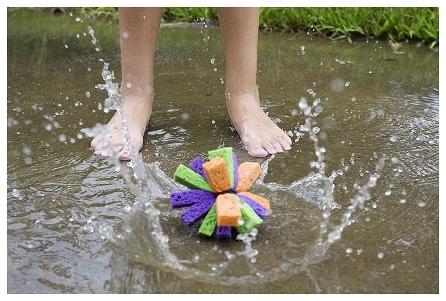 A purple, orange and green sponge bomb in a puddle of water. A child splashes around it. 